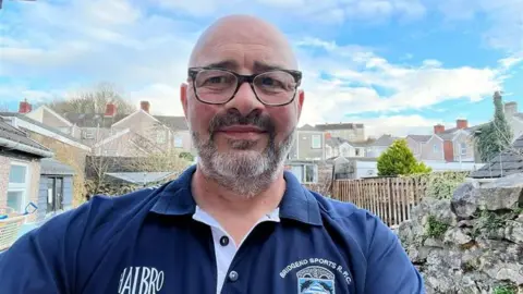 Chris Rees Selfie of Chris standing in his garden wearing a Bridgend RFC navy club t-shirt. He is bald, wears glasses and has a short grey beard.
