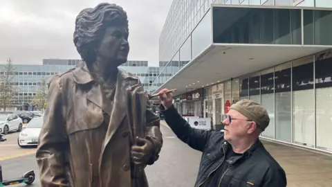 Amy Holmes/BBC The image shows a life‑sized statue of Jennie Lee MP standing outdoors beside artist Andy Edwards, who is touching the statue's shoulder while holding a cloth. The scene is set on a paved walkway next to a modern glass‑fronted building, with parked cars and electric scooters visible in the background under an overcast sky.
