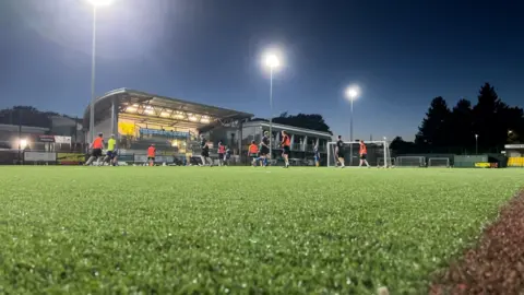 A dark astro turf where two teams are playing a match. One team is wearing navy shirts and the other is wearing orange bibs.