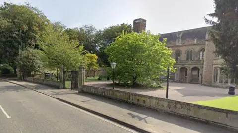 Google A stone-built, large hall sited next to a main road. Around the hall is a low wall. Trees and shrubs are in the background.