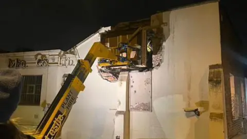 Phoebe Carys At night, a boom lift is used to remove part of an the building’s wall, revealing its interior. A worker in a high-vis jacket is inside. 