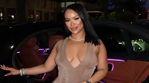 Instagram A woman in a light brown dress stands in front of a car, with a shell necklace and straight black hair.