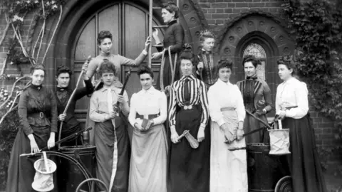 Courtesy of The Mistress and Fellows, Girton College, Cambridge A black and white image of 11 late Victorian Girton College students standing against a building. They are holding fire fighting equipment including two water pumps, hoses, rope, a rattle and one is standing up a ladder. They all have their hair up and are wearing long dresses. 