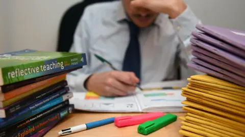 A teacher in a light coloured shirt and dark tie looking stressed while marking work. There are piles of txt books and pupils' work books stacked up on either side of his desk and there are pens and colourful markers in front of him.