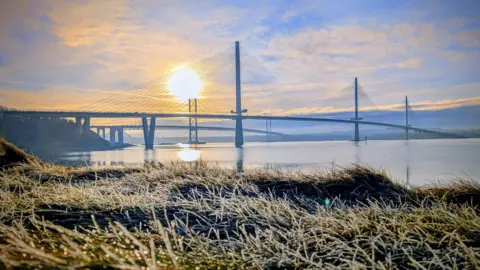 Laura McLean The morning sun rises behind a cable-stayed bridge, casting reflections on calm water, with frost-covered grass in the foreground.
