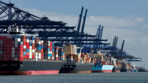 A row of ships filled with containers docked by a port filled with cranes