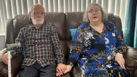 Paddy and Breda Murphy sitting on a brown sofa, holding hands. Paddy is holding a walking stick