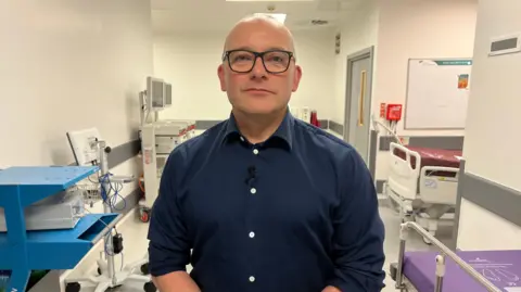 A man in his mid-fifties is stood in a hospital corridor.  He is wearing a dark blue shirt and is wearing black rimmed glasses.  There are different pieces of medical equipment in the corridor behind him, including a bed and some trolleys with computer monitors on them.