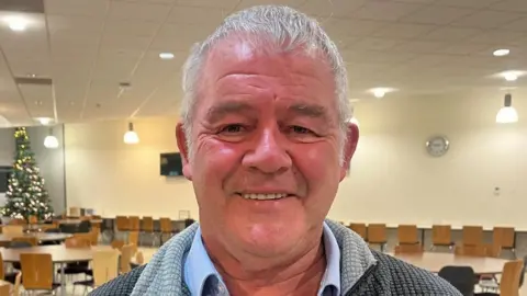 A man with short white hair wearing a waffled jumper and blue shirt stands in a rehearsal room with many wooden chairs and a Christmas tree in the distance.