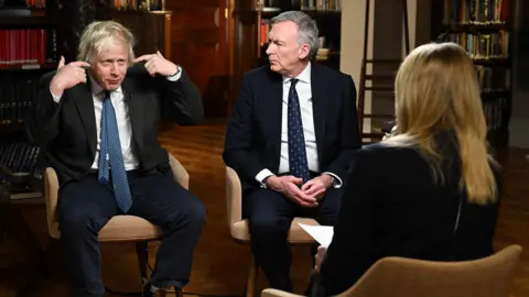 Former prime minister Boris Johnson speaking while pointing fingers at the sides of his head. A suited man with greying hair looks at him. In the foreground, out of focus, we see the back of a woman with blonde hair.