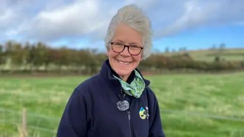 Denise Walton, who has grey hair and dark glasses, is wearing a blue zipped fleece and a colourful scarf. She is standing in a field with a long hedgerow behind her.