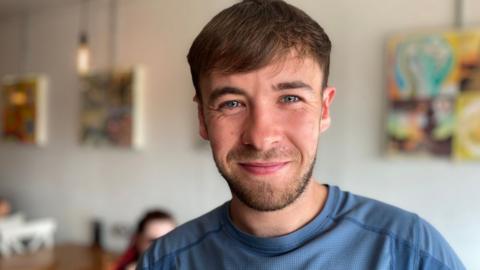 A smiling Lews Quail, with short brown hair, blue eyes and a close-cut beard, wearing a blue-grey T-shirt and standing in a white-walled cafe which has abstract multi-coloured artworks hung inside