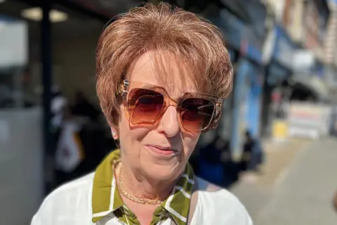 BBC Woman in white jumper, green shift and big sunglasses looks at camera outside a shop
