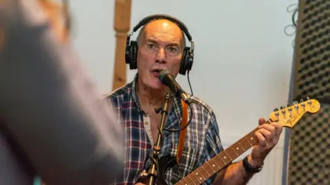 Dr Curran sings and plays the guitar in a studio. He has a serious expression and is looking away from the camera. He wears large black headphones and a muilt-coloured checked shirt. 