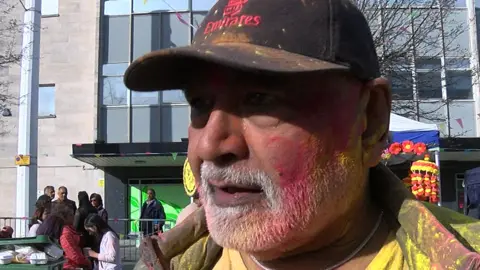 Avinas is wearing a black cap which says Fly Emirates on it. He is wearing a yellow T-shirt and a coat, both of which have been covered in yellow and pink powder. He has a white beard and part of his face has been covered in pink and yellow powder.