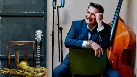 A man with dark brown hair and wearing a blue suit with a white shirt and blue tie sits on a green chair, learning his head on his hand. A large string instrument can be seen behind him, alongside a microphone stand, guitar and trumpet resting on a wooden chair.