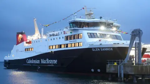 PA Media The Glen Sannox sits on the water. It is a white and black ferry with "Caledonian MacBrayne" written on the side. It is early morning and only just starting to get light, with heavy clouds in the sky