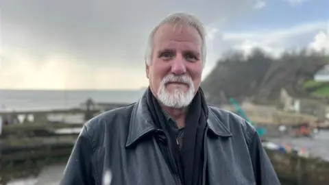 Rolf Munding smiles at the camera, he is wearing a black leather jacket and has a grey beard.
