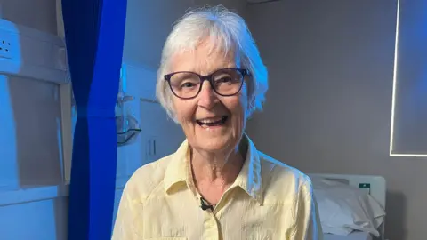 A woman with short white hair and glasses sits in a clinical setting, wearing a light yellow shirt with a small microphone clipped near the collar. The background includes a hospital bed with white bedding, a blue privacy curtain, medical wall fixtures, and a window with soft lighting entering from the side.
