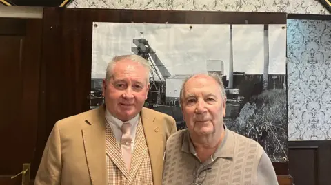 Steve Jones/BBC Johnny Wood poses for a photo with another former miner. Both men have short grey hair and brown eyes. They wear a neutral expression in the photo.