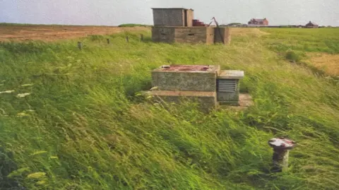 Carl Olsen A weathered, squat, rectangular concrete block, with a rusty metal hatch on top, stands in a field of overgrown green grass. A metal post stands to the right. In the background, a larger concrete building with small rectangular firing slits in the sides, can be seen.