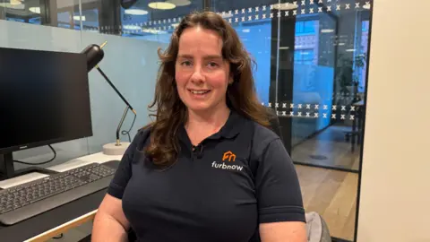 Becky Lane in a picture. She has curly hair and is smiling at the camera. She is sat in an office. There are glass panes behind her and a computer. She is wearing a navy blue polo shirt with the name of her firm on. 