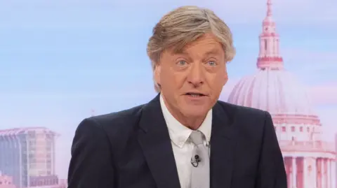Richard Madeley sitting behind a desk presenting Good Morning Britain. He is wearing a dark suit jacket, white shite and grey tie. Behind him the dome of St Paul's Cathedral can be seen.