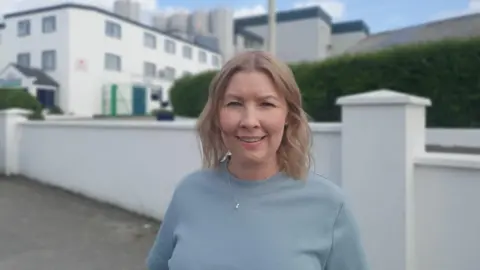 A women in a blue jumper stands in front of large white wall. She has light brown hair. a factory building can be seen behind the wall
