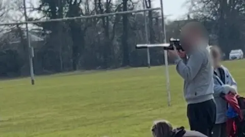 BBC A man in a grey jacket at a pitch side looking down a barrel-like fitting on the device on a football pitch. 