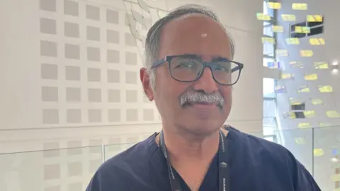 Anil Varma is standing in a large entrance hall, smiling at the camera. He has grey hair. He is wearing black glasses, and is wearing navy blue scrubs.