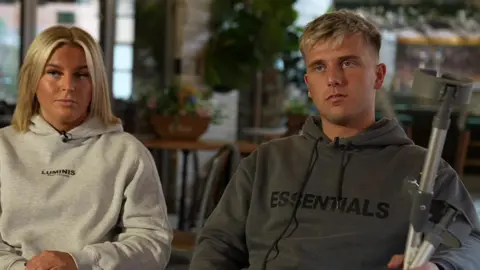Jack has blonde hair and is wearing a grey hoodie. He is holding crutches and is sitting down. Abbie has shoulder length blonde hair and is wearing a lighter grey hoodie. She is sitting beside him.