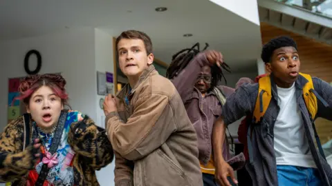 One young woman and three young men look startled by something they see off camera, looking as if they have been stopped in their tracks. All but one is carrying a backpack or bag. 