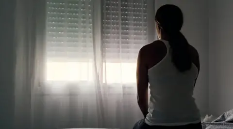 Getty Images A woman sat on a bed in a dark room looking at a shuttered window.