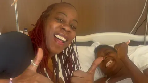 Handout Lavern Gilzene next to her daughter in the hospital bed. Both are smiling and making a thumbs up sign. 