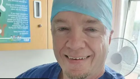 Robert Albon, who appears to be in a medical facility. He has a blue top, possibly a gown, that is just visible and a blue head covering as you might see in a surgical setting. He is smiling and has a small amount of short light coloured beard growth on the angle of his chin, roughly the width of his mouth.