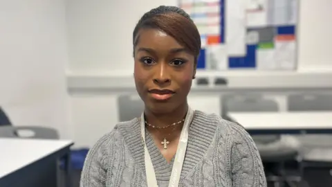 Head and shoulders of Donna Gezellekarikari who is wearing a grey jumper and lanyard. Grey chairs and a white table can be seen in the background.