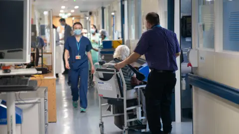 A hospital corridor with several healthcare workers in blue scrubs, some wearing face masks. In the foreground, a staff member pushes an elderly person with white hair in a chair or wheelchair, while other clinicians work in the background.