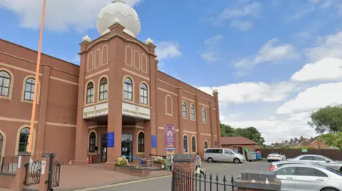 An orange brick building stands to the left of the image with a car park on the right. The building has has an extended entrance that stands out from the rest of the building with columns around the front door. There are arched windows on the top and bottom levels and the name of the temple in Punjabi and English above the entrance.