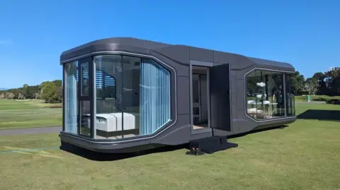 Capsule Holiday Homes/ Alice Gregory A sleek, modern modular cabin with dark grey exterior panels and large floor-to-ceiling glass windows sits on a grassy field under a clear blue sky. The structure has rounded corners, a minimalist design, and an open door revealing a clean, contemporary interior with a bed visible through the glass.