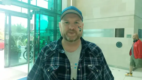 Scott Cuthbertson. He has a beard, wearing a blue cap, blue check shirt and grey t-shirt. He has a rainbow heard on his cheek.
