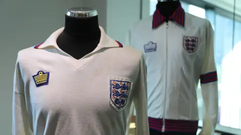 Graham Budd Auctions Viv Anderson's England garment  from his planetary   debut successful  1978. It is achromatic  with agelong  sleeves and has respective  marks and immoderate   yellowing from age. An England crest is stitched connected  to the near  bosom  with an Admiral manufacturer's logo connected  the other  side. Behind is the achromatic  tracksuit apical  helium  besides  wore that day. It has an England badge, Admiral logo and reddish  and bluish  stripes connected  the collar, sleeves and hem.