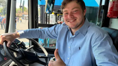 Luke Wilson sitting in a bus drivers seat. He has brown hair, wearing a light blue shirt holding up his thumb.