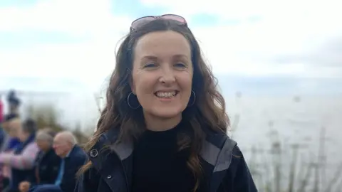 Una smiling at the camera. She has long wavy brown hair and is wearing sunglasses on top of her head, a black top and jacket and hoop earrings. 