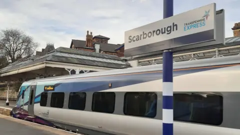 A railway station sign reading "Scarborough" with a train in the background standing at a platform.