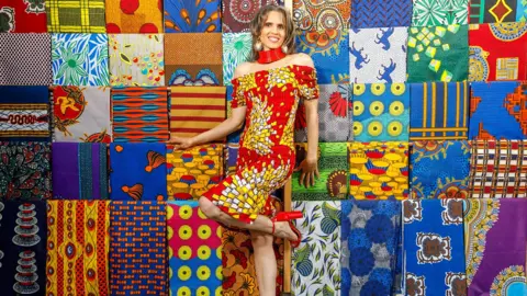 Darren Watley A woman with long brown hair stood in front of a multi-coloured quilted display. She is wearing an off-the-shoulder red dress, with a white and yellow graphic pattern, and red high heels. She has a thick red necklace and large earrings. She is posed with one knee raised and her hands touching the quilt.