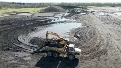 A lorry and a yellow digger at work on the muddy site of the new hospital. There are large puddles.