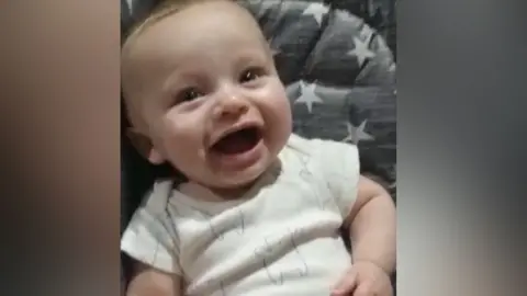 Police handout A smiling baby sitting in a grey and white star-patterned highchair in a white vest