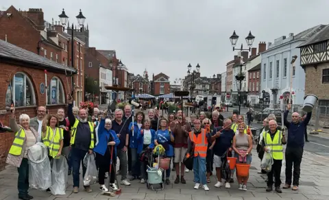 Carmel Wilson About 30 people, some in high-vis jackets, pose for the camera in the foreground. Many are carrying cleaning equipment. Buildings are in the background.
