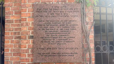 Jack Hadaway-Weller/BBC A stone plaque on the wall of a car park in both Hebrew and English.