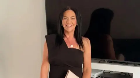 Karen Cummings, who has long dark hair parted in the middle, smiles while wearing a black dress and silver necklace and standing in front of a large flat screen TV 